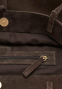 Interior de un bolso de ante marrón con un bolsillo de cremallera negra. Cuenta con un cierre de botón de latón y detalles de costura. Textura suave y diseño minimalista.