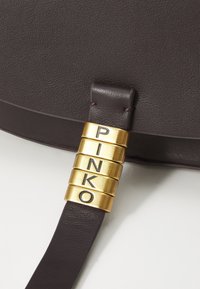 Καφέ δερμάτινη θήκη τσάντας με χρυσή μεταλλική λεπτομέρεια που φέρει χαραγμένα τα γράμματα "PINKO." Λείος υφασμάτινος texture και μινιμαλιστικό σχέδιο.