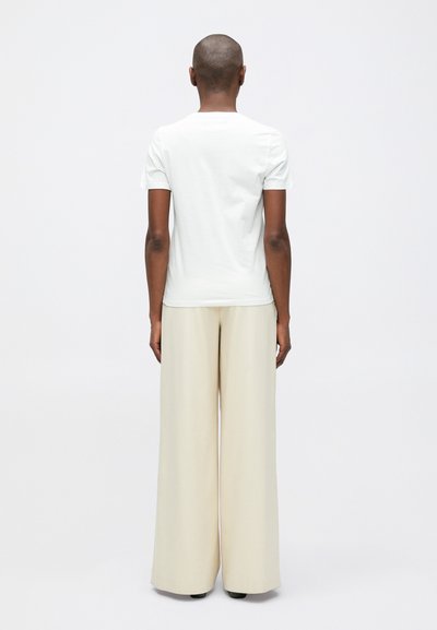 Osoba s oholenou hlavou, oblečená v jednoduchém bílém tričku s krátkým rukávem a širokých béžových kalhotách, stojící na bílém pozadí.