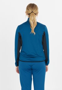 Sininen urheiluotsa mustilla yksityiskohdilla, jossa on korkea kaulus ja pitkät hihat. Tasainen pinta, tiukka malli, yhdistettynä sinisiin urheiluhousuihin.