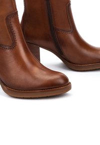 Botas de cuero marrón hasta el tobillo con puntera afilada, cremallera lateral y tacón apilado. Textura suave y detalle de costura en la parte superior.