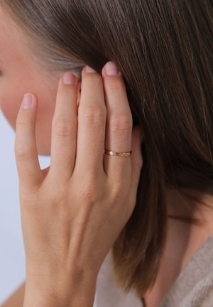 Goldfarbener Ring mit strukturiertem Design, der an einer Hand getragen wird, während die Finger die Haare berühren. Glatte Hauttextur und ein natürliches Nagelbild.