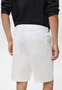 Pantalones cortos de lino blancos con corte recto, que cuentan con dos bolsillos traseros. Llevados con una camisa negra de manga larga, mostrados desde la parte de atrás.