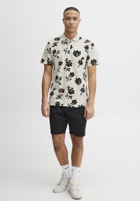 Kortermet polo i kremfarge med svart blomstermønster, matchet med svarte shorts og hvite joggesko. Har tre knapper på halsen og smal passform.