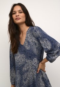 Mörkblå klänning med paisley-mönster och vita detaljer. Har en V-ringning och puffärmar, tillverkad av ett lätt, strukturerat tyg.