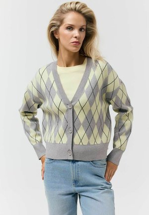 Kobieta ubrana w szary i bladożółty kardigan w romby na guziki, na jasnżółtej bluzce, w jasnoniebieskich dżinsach, stojąca z rękami na biodrach.