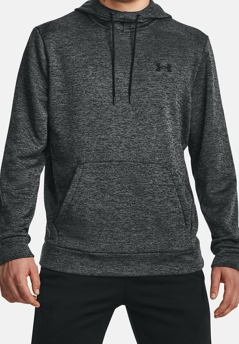 Homme portant un sweat à capuche gris foncé chiné avec poche kangourou à l'avant et cordons noirs, associé à un pantalon noir, debout sur fond blanc.