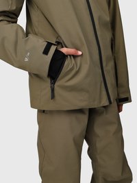 Persona che indossa una giacca impermeabile verde oliva e pantaloni abbinati, con una mano nella tasca con zip della giacca, manica interna nera visibile.