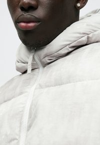 Gai pelēks zip-up pūkains jaka ar tekstūrētu apdari, aprīkota ar kapuci un maigu spīdumu. Aprīkojums ietver centrālo rāvējslēdzēju un regulējamas auklas.