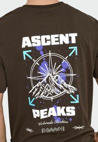 Brun bomulds t-shirt med et grafik af bjerge og et kompas, med hvid, blå og lilla sprøjtetilbehør og tekst der siger "ASCENT PEAKS."
