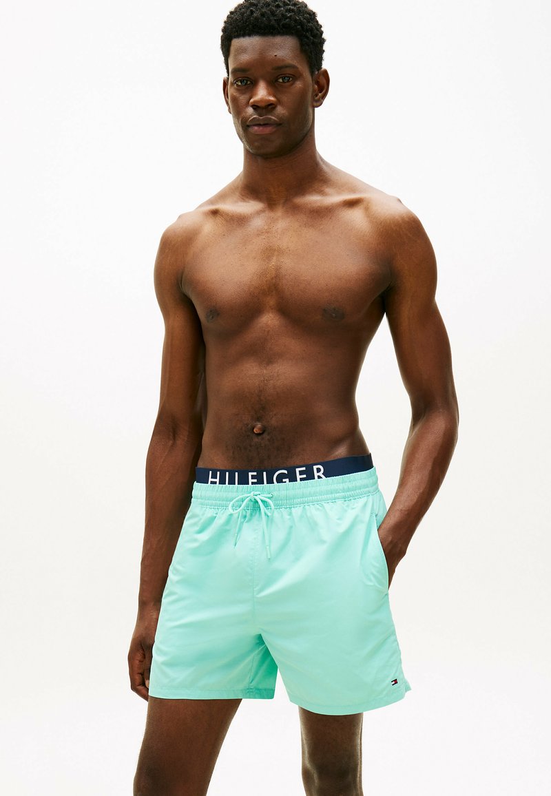 Homme torse nu portant un short de bain bleu clair avec une ceinture bleu marine arborant le texte "HILFIGER", debout devant un fond blanc uni.