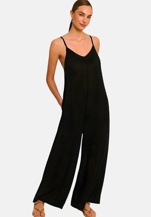 Femme portant une combinaison noire ample sans manches avec des bretelles fines et des jambes larges, associée à des sandales marron ouvertes à l’avant, debout les mains dans les poches.