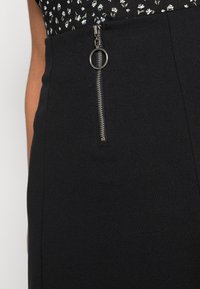 Jupe noire avec une fermeture éclair verticale munie d'un tirette circulaire, confectionnée dans un tissu lisse et texturé. La ceinture est ajustée et plate.