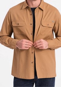 Brązowa koszula zapinana na guziki, wykonana z teksturowanej tkaniny, z dwoma kieszeniami na piersi i kołnierzykiem. Nosi się ją na czarnej warstwie pod spodem.