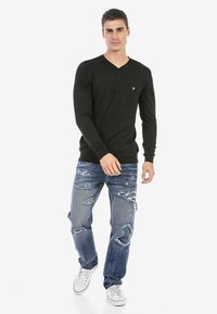 Schwarzer Langarm-V-Ausschnitt-Pullover mit kleinem Emblem, kombiniert mit ausgewaschenen blauen Jeans und weißen Sneakers mit roten Akzenten.