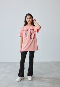 Pink T-shirt med stort "TB" grafik, kombineret med sorte flared bukser med "BAKER" i pink på underbenet. HvIde sneakers fuldender looket.