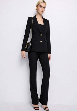 Donna che indossa un blazer nero su misura con bottoni dorati e pantaloni neri abbinati, mentre tiene una borsa nera con tracolla a catena dorata.