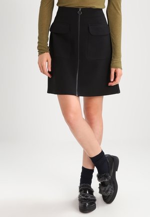 Kvinde iført sort nederdel med frontlynlås og lommer, olivengrøn langærmet top, sorte sokker og sorte loafers med pelsdetaljer.