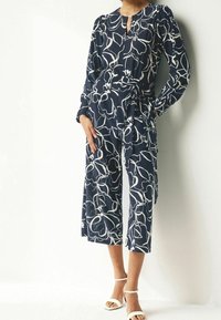 Combinaison bleu marine avec un motif floral blanc, manches longues, décolleté en forme de clé, taille ceinturée et design à jambes larges cropped. Portée avec des talons blancs.