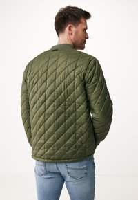 Olivově zelená prošívaná bunda s texturovaným povrchem, kulatým límcem a žebrovanými manžetami, nošená s modrými džíny. Zadní pohled ukazuje diagonální prošívání.