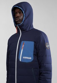 Giubbotto imbottito blu navy con una tasca sul petto bicolore blu, zip bianca e cordino arancione; presenta un cappuccio aderente e un marchio sulla manica.