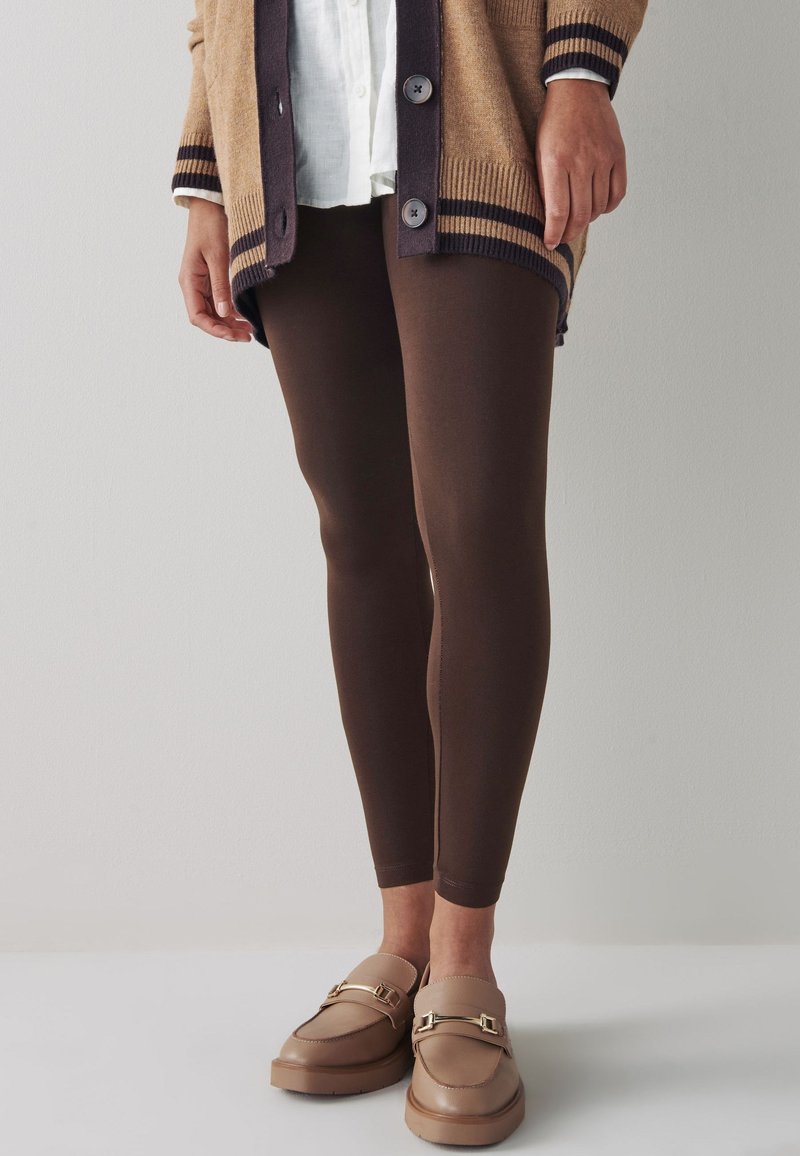 Braune Leggings aus elastischem Stoff, kombiniert mit einer beigen Strickjacke mit dunklen Streifen und Knöpfen sowie hellbraunen Slip-on-Schuhen mit Hardware-Akzent.