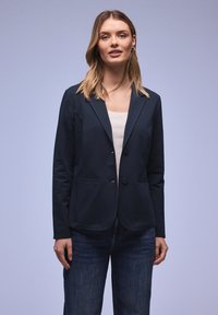 Femme aux cheveux blonds portant un blazer bleu marine sur un haut blanc et un jean bleu, debout devant un fond uni bleu clair.