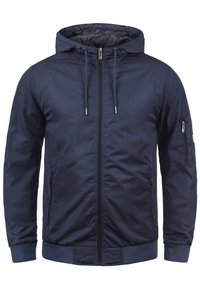 Veste légère à capuche bleu marine avec fermeture éclair frontale, capuche réglable avec cordon, poches latérales zippées et poche zippée sur la manche.