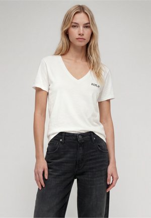 Blonde Frau mit weißem V-Ausschnitt-T-Shirt mit "Replay" Logo und schwarzen Jeans, stehend vor einem einfarbigen hellen Hintergrund.