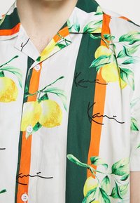 Camisa de manga corta con estampado de limones, rayas verdes y naranjas, botones blancos y detalles bordados en negro.