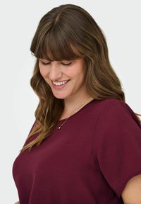 Burgundy kortärmad topp med rund halsringning, i en texturerad tygkvalitet. Ett delikat guldhalsband med en rund medaljong syns.