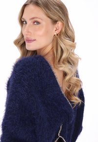 Marineblauwe fuzzy trui met een V-halsontwerp, versierd met zilveren bies. Model heeft golvend blond haar en draagt gouden studoorbellen.