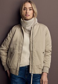 Beige bomberjack met een gladde buitenkant, ribgebreide manchetten en een rits. Gelaagd met een lichtbeige gebreide coltrui. Donkere jeans maken de outfit af.