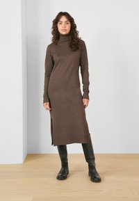 Robe brune longue à col roulé en tissu tricoté doux, dotée d'une fente sur le côté, associée à des bottes noires à hauteur de genoux. Design simple et ajusté.