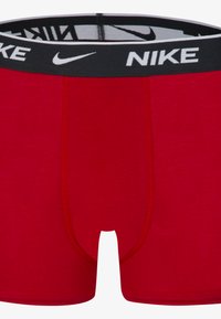 Rde bombažne bokserice z črnim pasom, na katerem je v beli barvi logotip Nike. Gladka tekstura in prilegajoč dizajn s šivi spredaj.