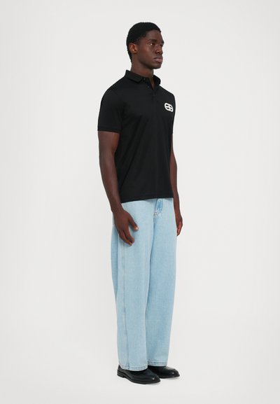 Bărbat stând în picioare, îmbrăcat cu un tricou polo negru cu logo alb, blugi largi de culoare albastră deschisă și pantofi negri, pe un fundal alb simplu.