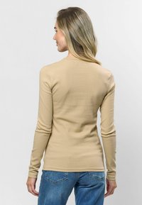 bézs hosszú ujjú felső bordázott textúrával, kerek nyakkal és testhez simuló fazonnal. Kék farmerral párosítva, hátulról nézve.