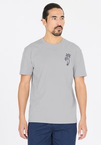 Harmaa puuvillapaita, jossa on lyhyet hihat ja pyöreä kaula-aukko, vasemmassa rinnassa pieni laivastonsininen palmupuu -grafiikka.