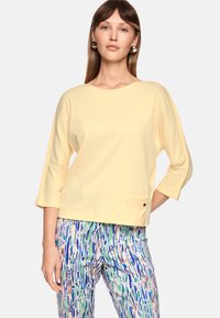Yellow, 3/4 sleeve top with a round neckline and a front pocket. Soft fabric with a relaxed fit. Paired with patterned, multicolored pants.