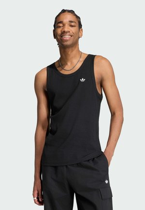 Giovane uomo con capelli intrecciati che sorride, indossa una canotta nera e pantaloni cargo neri con loghi Adidas su entrambi i capi.