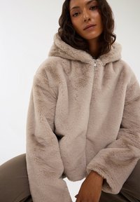 Giacca con cappuccio in pelliccia sintetica beige, con chiusura a zip, maniche oversize e texture morbida, caratterizzata da una vestibilità rilassata e tessuto soffice.