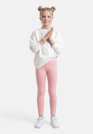 Young girl with braided hair in two buns, wearing white sweatshirt, pink leggings, and white sneakers, standing with hands clasped.
