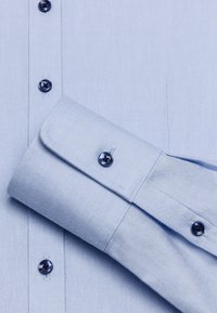 Camicia azzurro chiaro realizzata in tessuto con una sottile texture, caratterizzata da bottoni scuri e un polsino arrotolato con dettagli coordinati.
