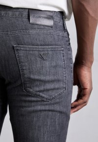 Grå denimjeans har en bakficka med en broderad logotyp och en läderetikett vid midjan. Tyget har en slät textur.
