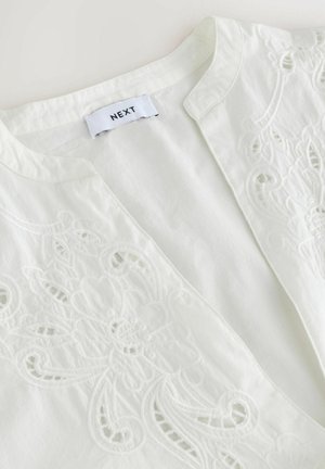 White blouse with intricate floral embroidery on one side, featuring a mandarin collar and smooth, lightweight material. Brand label visible.