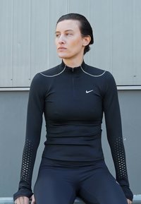Czarna sportowa bluzka z długim rękawem, z wysokim kołnierzem, z zapięciem na zamek i perforowanymi detalami na rękawach. Wykonana z dopasowanego, elastycznego materiału.
