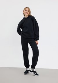 Svart hoodie och matchande byxor i mjukt material, med en avslappnad passform och ribbade mudd, ihop med svarta sneakers med vita detaljer.