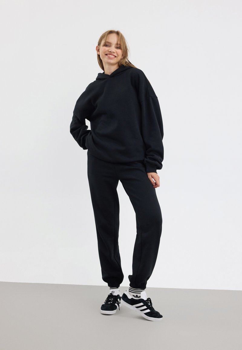 Svart hoodie och matchande byxor i mjukt material, med en avslappnad passform och ribbade mudd, ihop med svarta sneakers med vita detaljer.