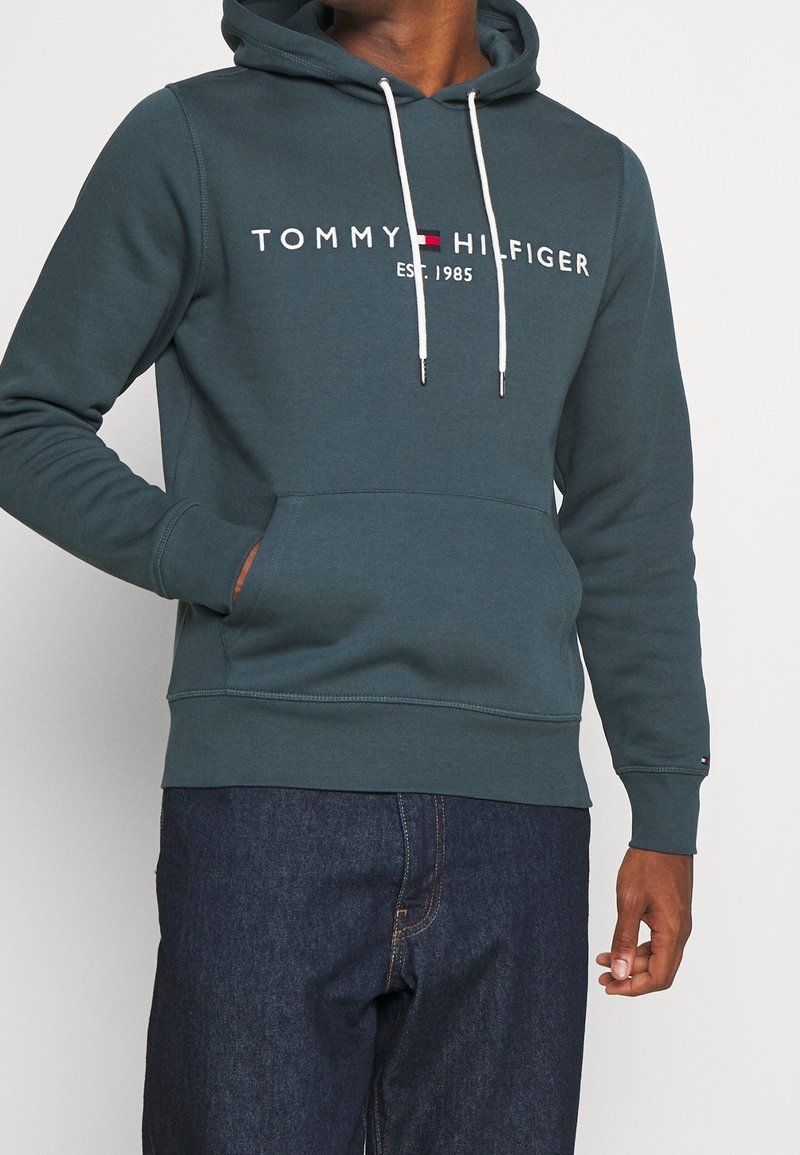 Dark teal hoodie with a kangaroo pocket, white drawstrings, and front logo "TOMMY HILFIGER EST. 1985." Features ribbed cuffs and hem.