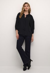 Blusa nera ampia con scollatura rotonda, dettaglio annodato e maniche svasate. Abbinata a jeans svasati in denim scuro e stivaletti neri.